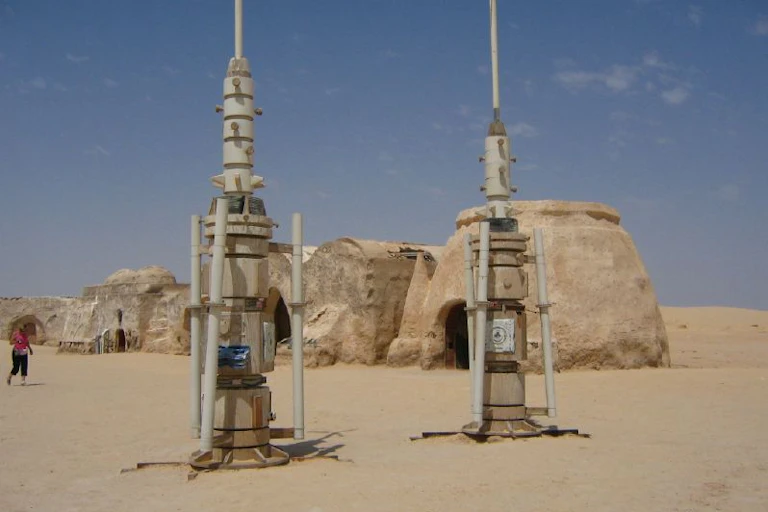 Two technological-looking towers stand in a desert landscape, with dome-like buildings behind them.