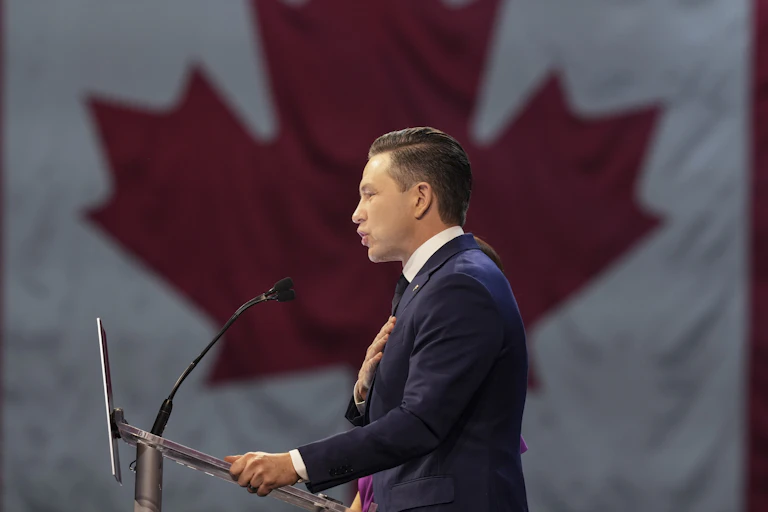 A man with dark hair on a stage with a large Canadian flag beside him.