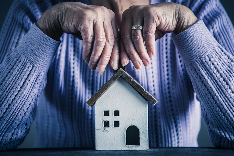 An elderly pair of hands above a small model of a house