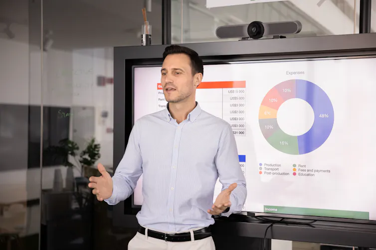 A man in a suit speaks to people off camera while standing in front of a pie chart