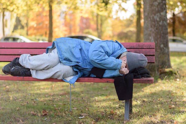 Elderly woman, in a grey hat, blue jacket and white pants, lies on a red park bench