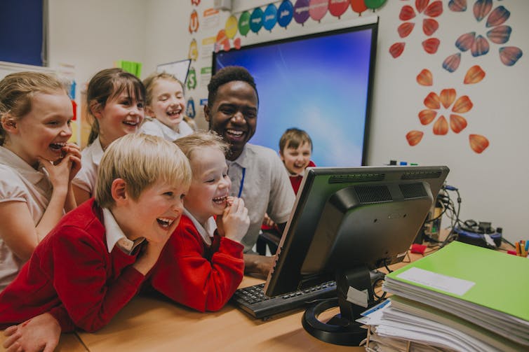 A group of primary students and a teacher crowd around a computer monitor, smiling and laughing.