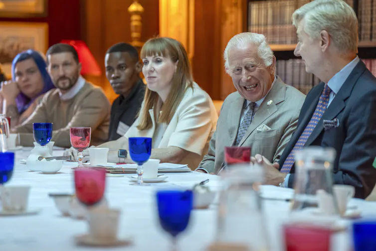 A man with grey hair smiles as he sits at a table with several other people. Red and blue water glasses are in front of them.