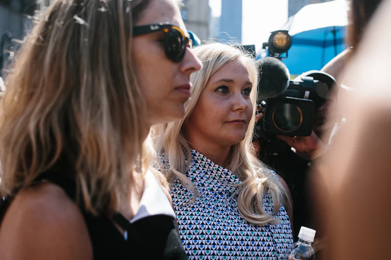Virginia Giuffre, with long blonde hair and a patterned dress, filmed by journalists outside of a courthouse with other Epstein survivors