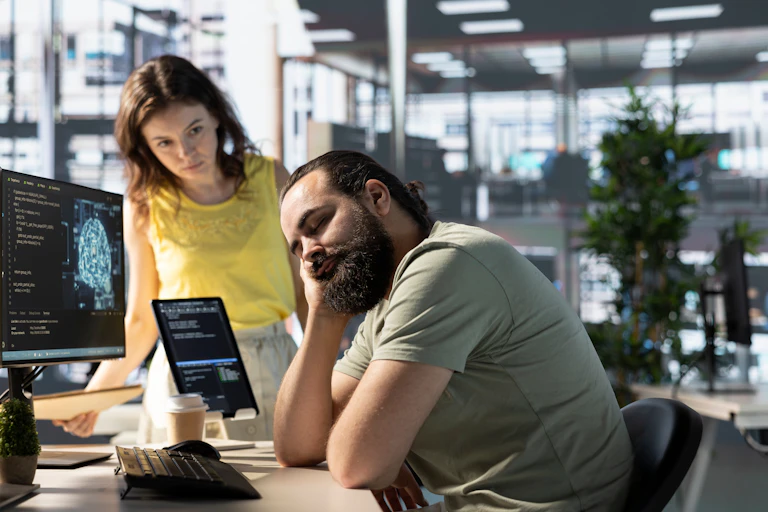 Une femme avec un Tshirt jaune regarde un homme barbu portant un t shit vert, la tête dans la main et les yeux fermés. IL est assis à son bureau, devant l'écan allumé de son ordinateur.