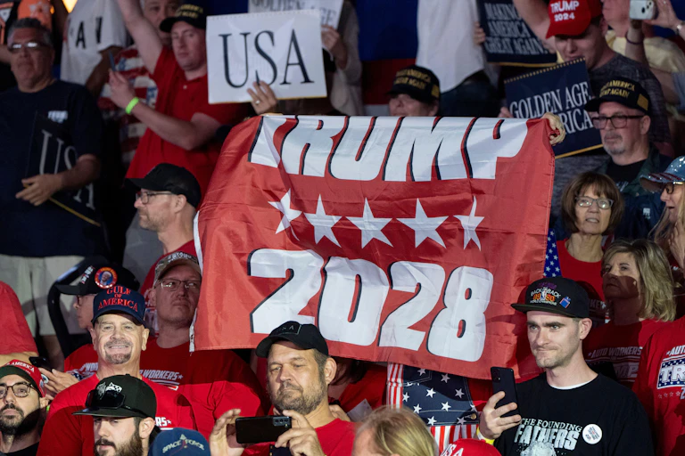 A person holding up a 'Trump 2028' flag in the middle of a crowd.