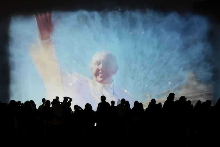 A large, slightly blurry image of a waving man in a white robe is projected over the silhouette of a crowd of people.