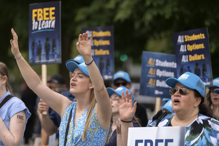 Two women raise their hands as though praying as they stand amid protesters wearing blue hats.