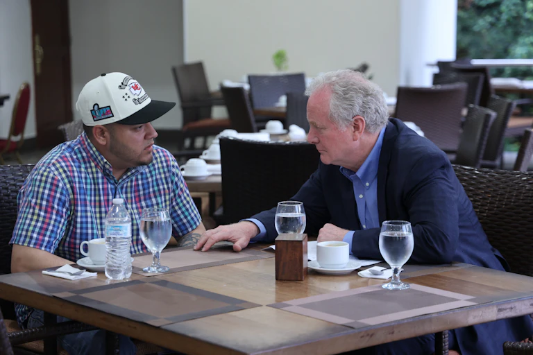 Two men sit together at a table - one wears a black blazer and the other wears a checkered button-down shirt and a white hat.