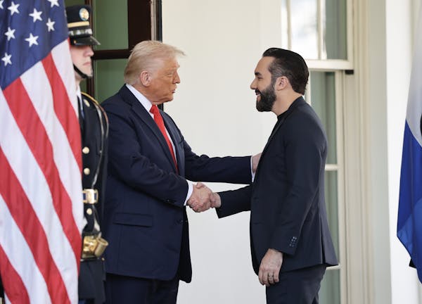 Two men in dark jackets shake hands next to a large American flag and a soldier standing next to it.