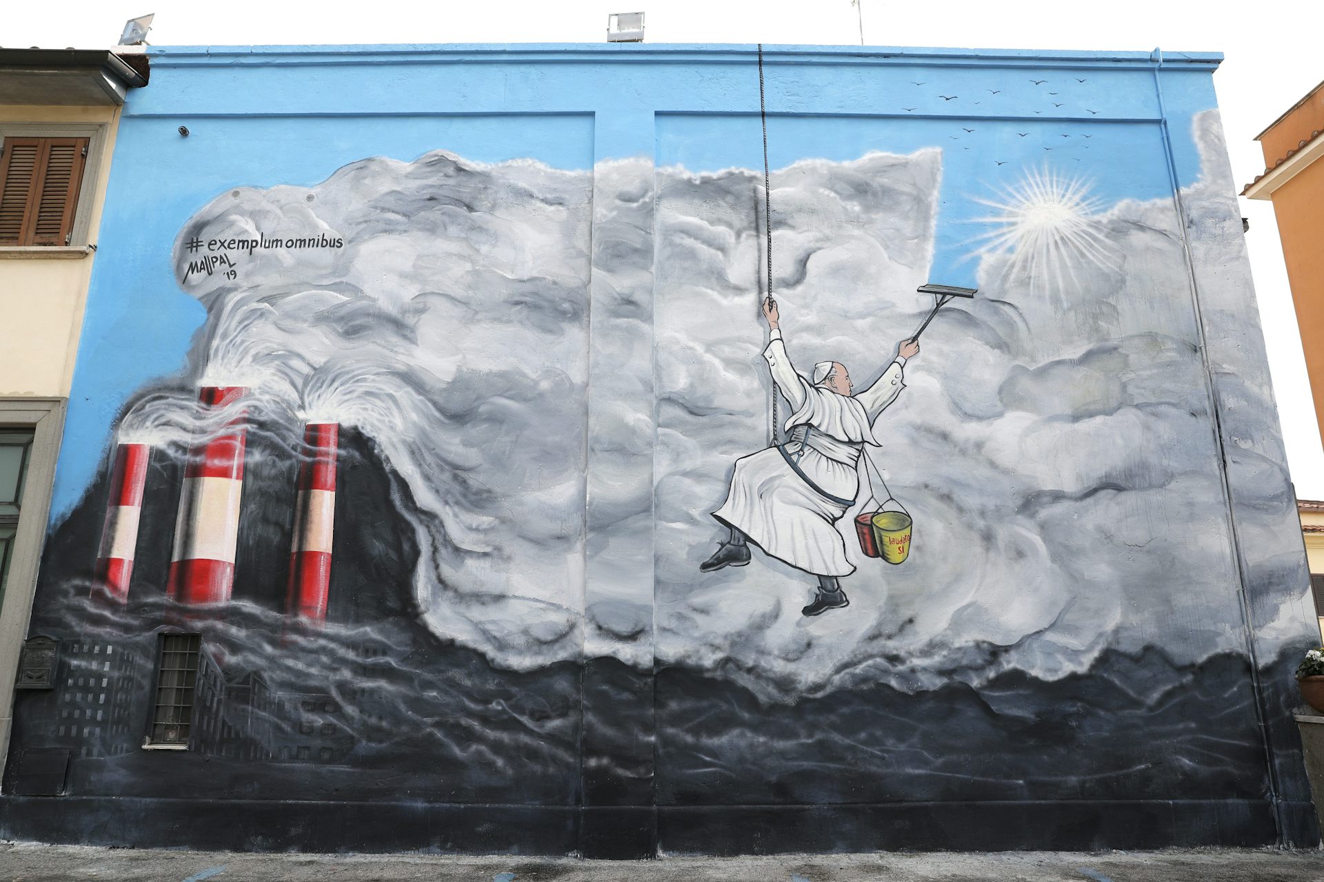 A large mural n a wall depicting a man in a white cassock wiping away smog emanating from smoke stacks to reveal a blue sky
