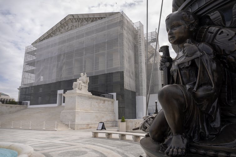 A grand building with large pillars is sheathed in construction scaffolding, with a statue of a child in the foreground.