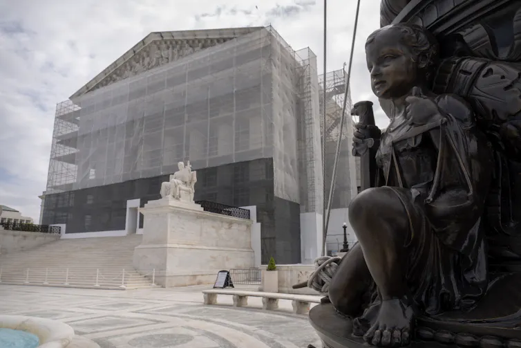 A grand building with large pillars is sheathed in construction scaffolding, with a statue of a child in the foreground.