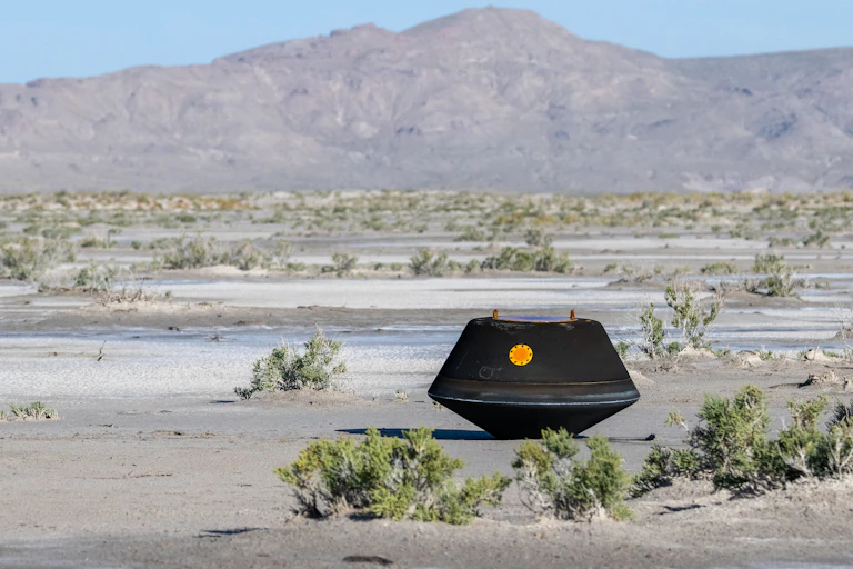 A sample return capsule, which looks like a box about the size of a microwave, sitting in a desert with a ridge in the background.
