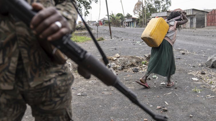 La lucha en la República Democrática del Este del Congo se deteriora a la violencia de género contra las mujeres 1 Una niña que usa jericano en la espalda pases por un hombre con neumáticos militares que usan rifle de ataque