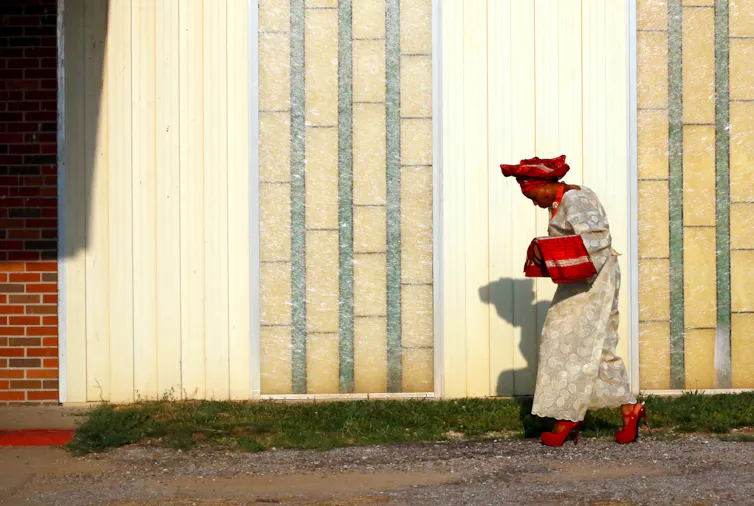 A woman wearing a bright red headscarf and red shoes walks near a building.