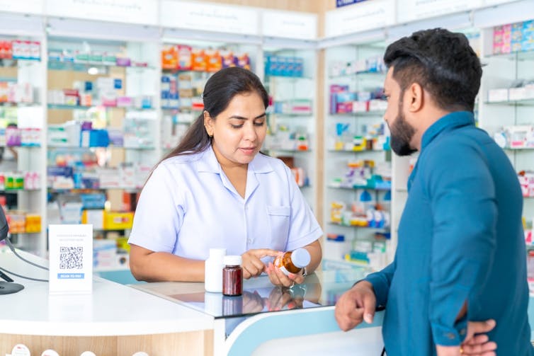 Man at pharmacy counter, female pharmacist checking medication label