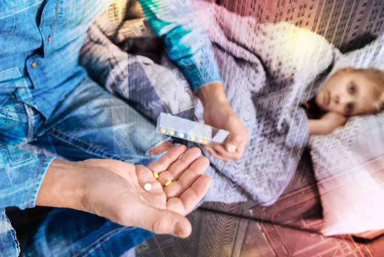 Father sitting at edge of sofa, holding medicine, while son lies on sofa under blanket