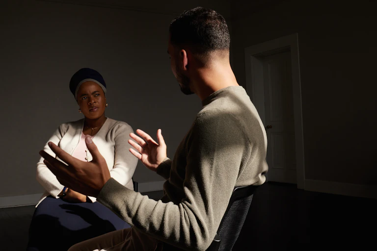 Two people sit in a dimly lit room talking.