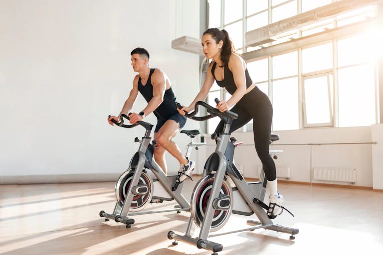 A young man and a young woman in workout gear cycle indoors on stationary bikes.