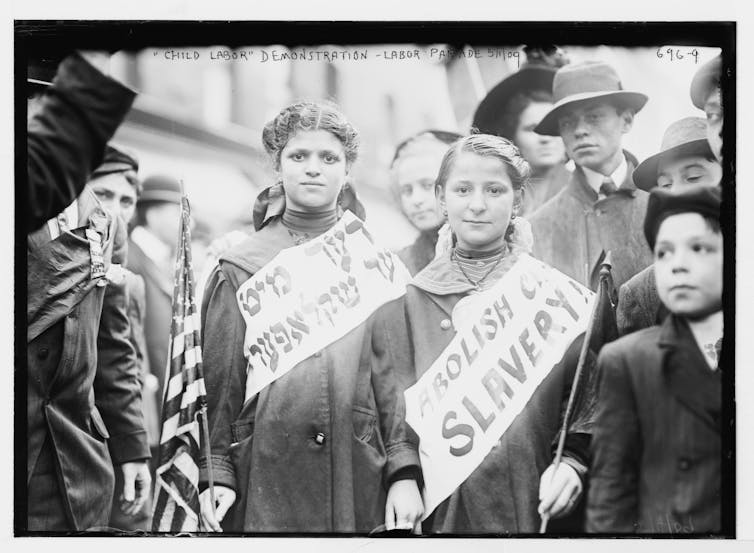 Two girls stand among group of people. One on left has sign in Yiddish. One on right has sign that says: Abolish slavery