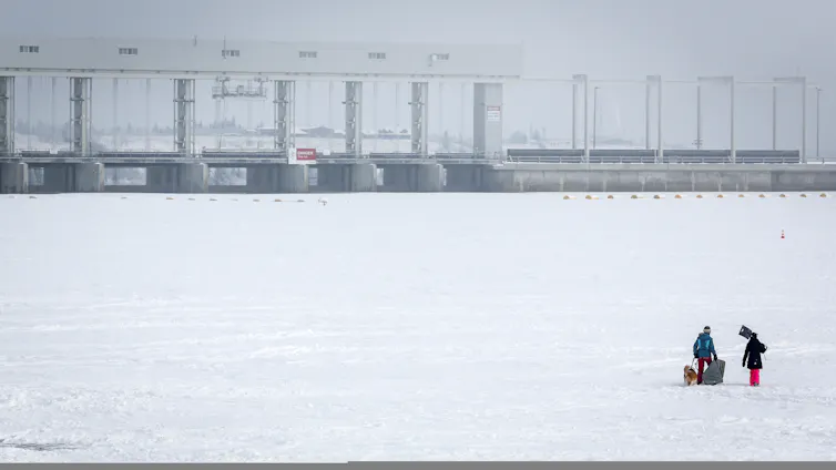 Dos personas limpian hielo en un lago congelado con una planta hidroeléctrica en el fondo.