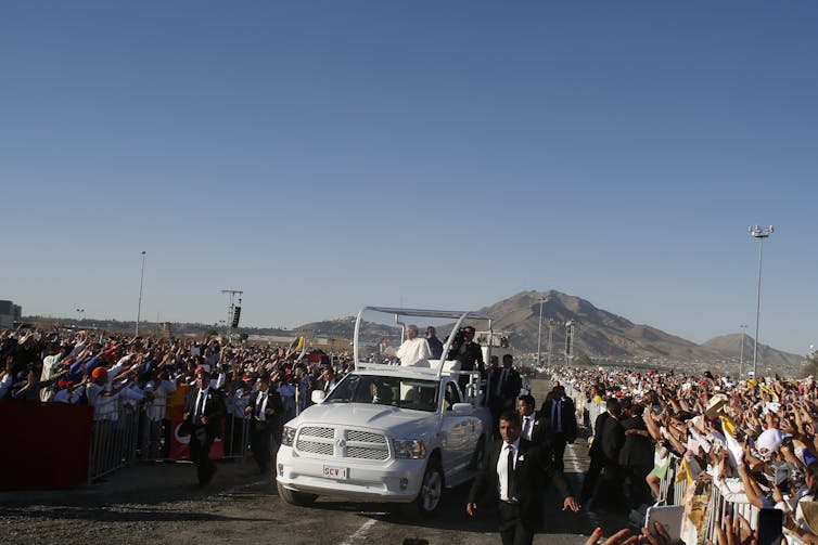 A white truck with people standing in its bed makes its way through a crowd outside, with a mountain visible in the distance.