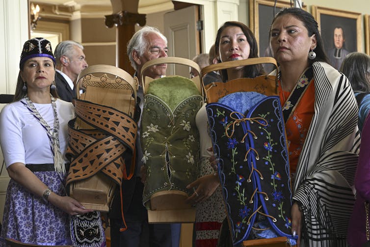 A man and three women stand holding embroidered cradleboards for infants.