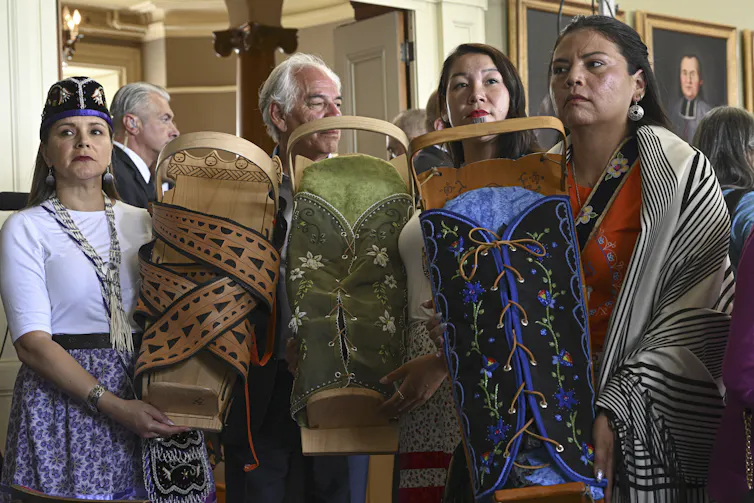 A man and three women stand holding embroidered cradleboards for infants.