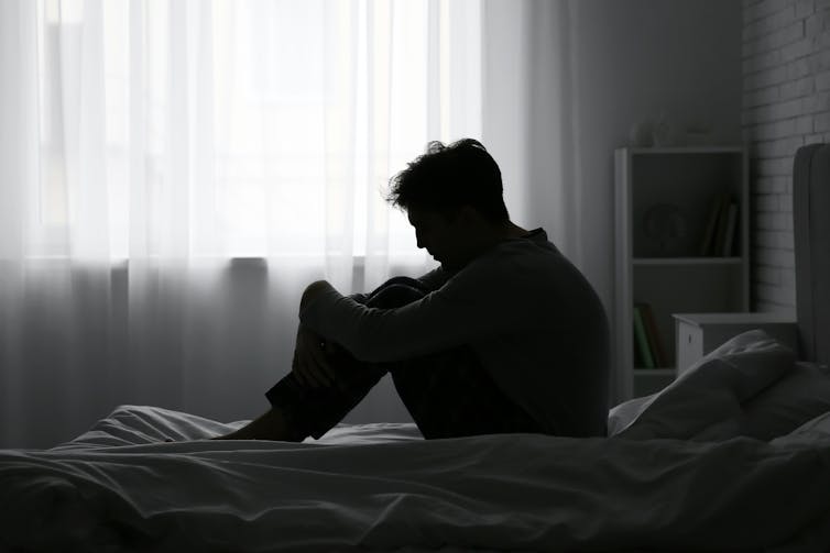 Silhoutte of man hunched in bedroom on bed.