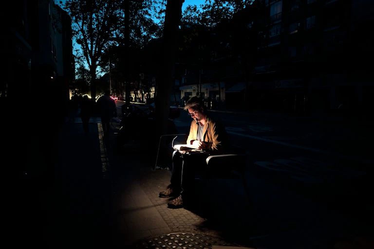 Un hombre leyendo en un banco en la calle alumbrado por una linterna.