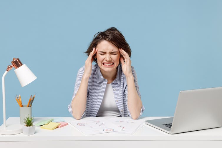 A stressed woman sitting at a desk, open laptop to the side.