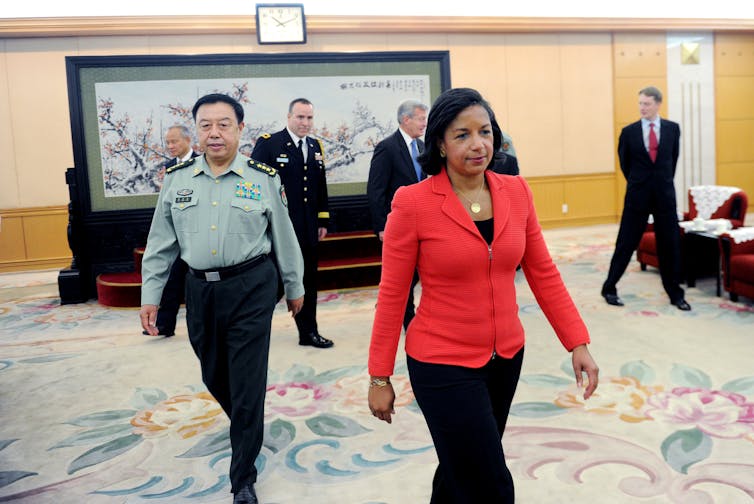 A woman dressed in a red top walks in front of a man dressed in military garments.