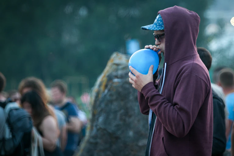 A man, outdoors and at a public gathering, is inhaling through a balloon.