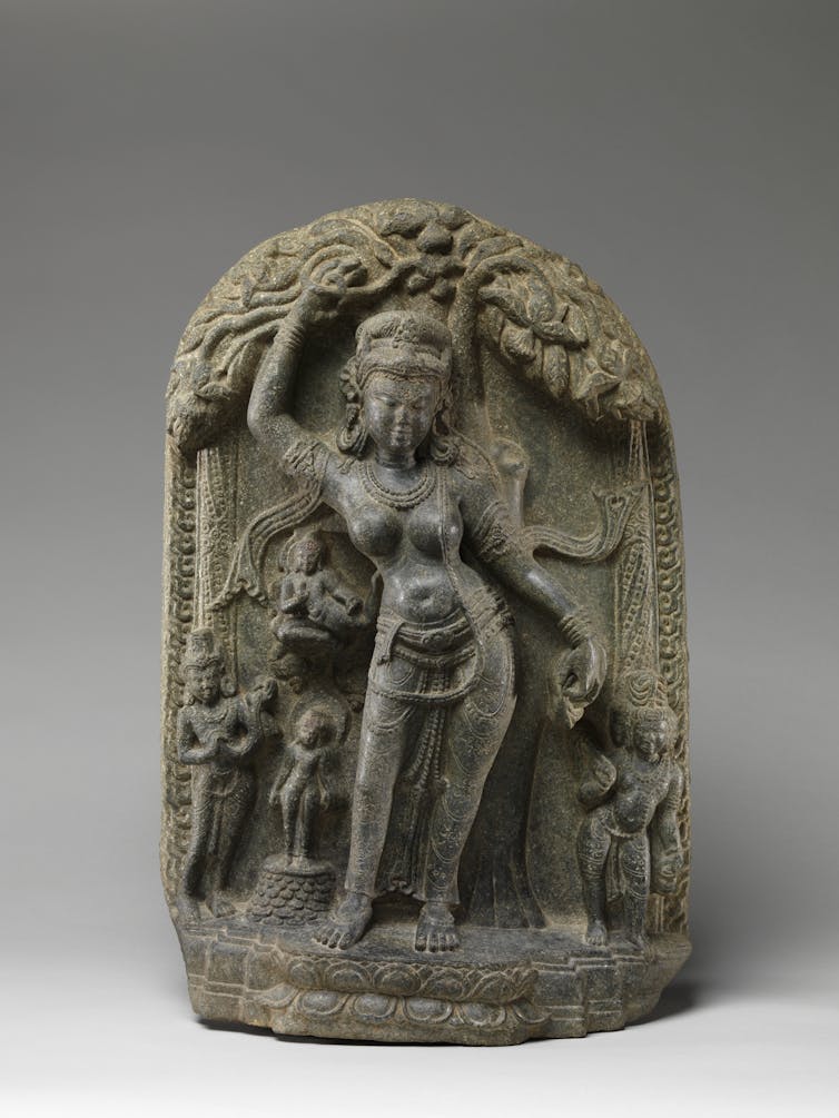 A statue of a woman with one arm extended above her holding the branch of a tree. On her right is a newborn standing on flower petals.