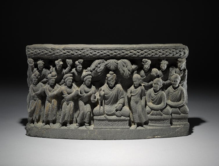 A panel showing a seated Buddha, with several figures on both sides. Prominent among those is a woman on his right offering a long cylindrical and tapering object like a flask.