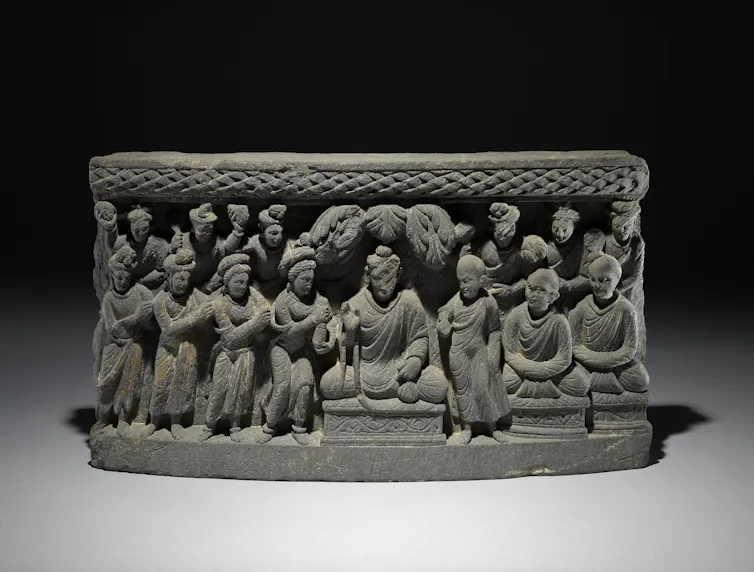 A panel showing a seated Buddha, with several figures on both sides. Prominent among those is a woman on his right offering a long cylindrical and tapering object like a flask.
