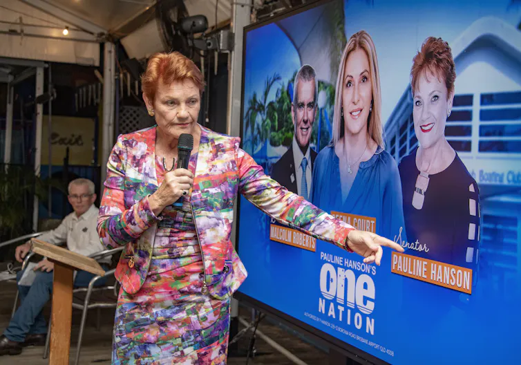 Red headed woman in colourful, patterned suit pointing at a bright blue corflute of herself