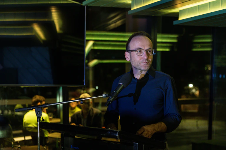 Glum looking man wearing a dark shirt, standing at a podium in a dark room