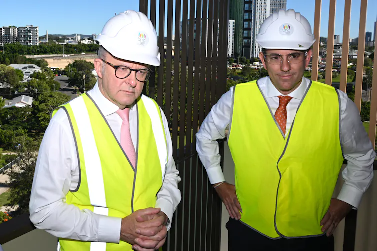 Anthony Albanese and Jim Chalmers wearing bright yellow hi-vis jackets and hard hats at a construction site.