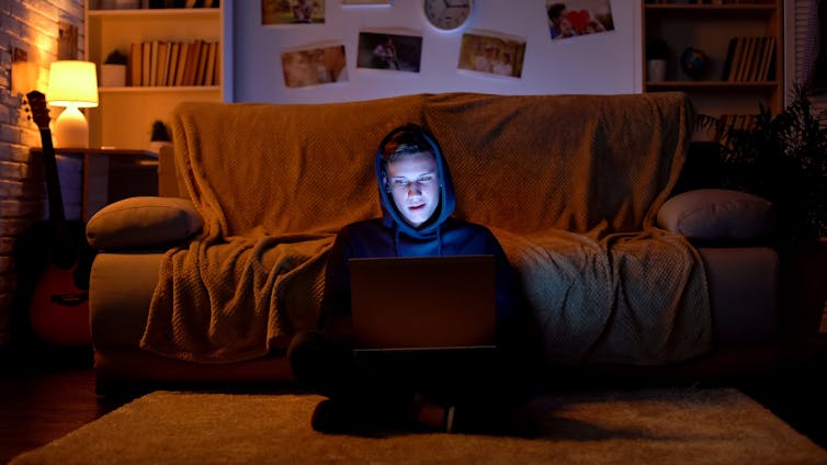 Boy in hoodie using laptop in a darkened room.