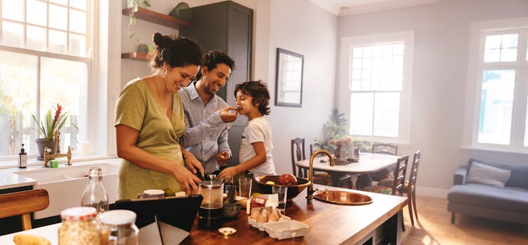 A man, woman and child chat and laugh in a kitchen while preparing food.