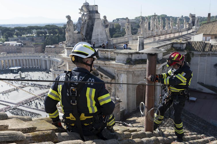 Dois bombeiros em um telhado com vista para a Praça de São Pedro.