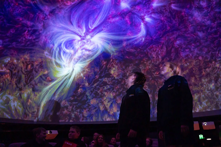 People stand in a dark room looking up at a ceiling with swirling purple nebulas.
