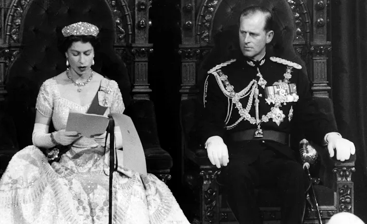 A black-and-white photo shows a woman with short dark hair in an ornate gown and wearing a crown reading a speech. A man in military garb sits next to her.