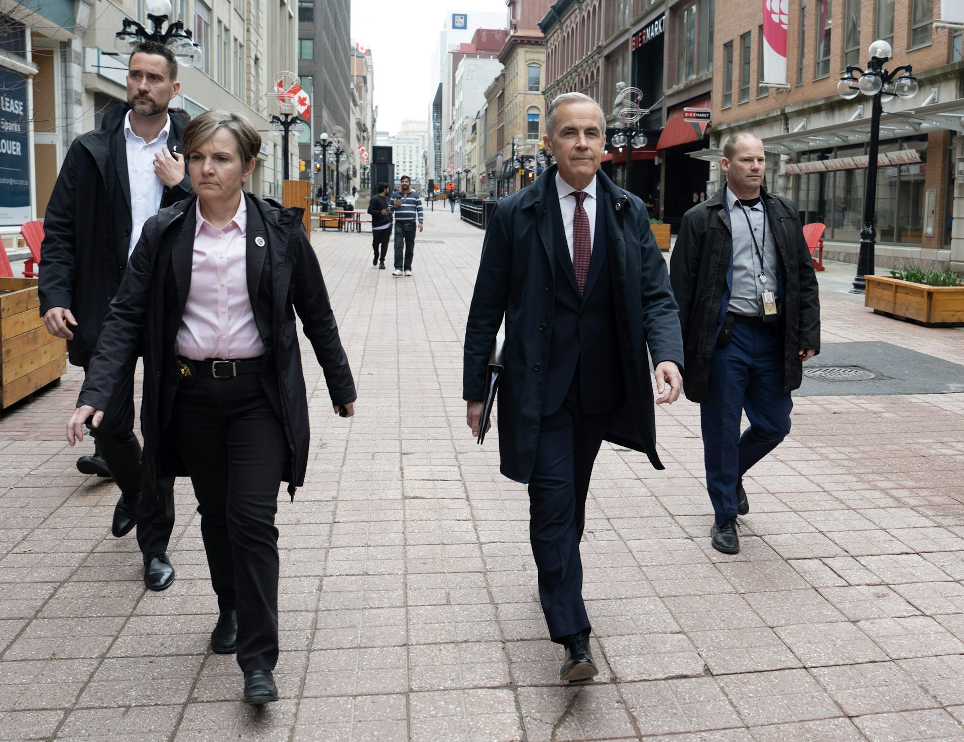 A man with short grey hair wearing an overcoat walks on a city walkway with people walking beside and behind him.