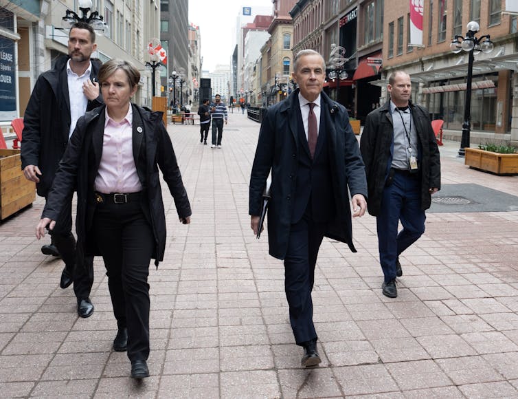 Un hombre con un cabello corto gris que lleva a California caminando en una ciudad caminando con gente caminando a su lado y detrás de él.