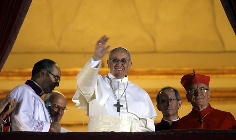 A man in a white robe with a large, simple cross around his neck waves and smiles from an open doorway.