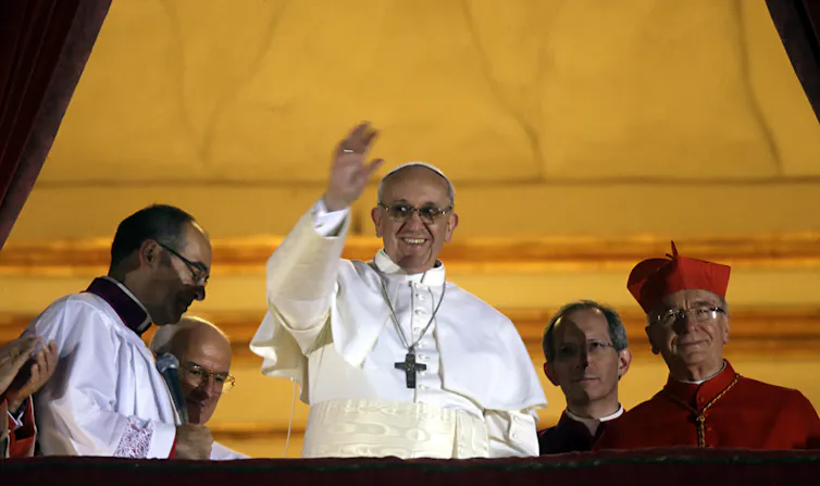 The pope in a white robe with a large, simple cross around his neck waves and smiles from an open doorway.