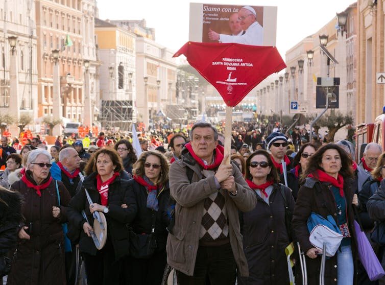 A group of people wearing red scarves, led by a man holding a picture of the pope, walk down a crowded street.